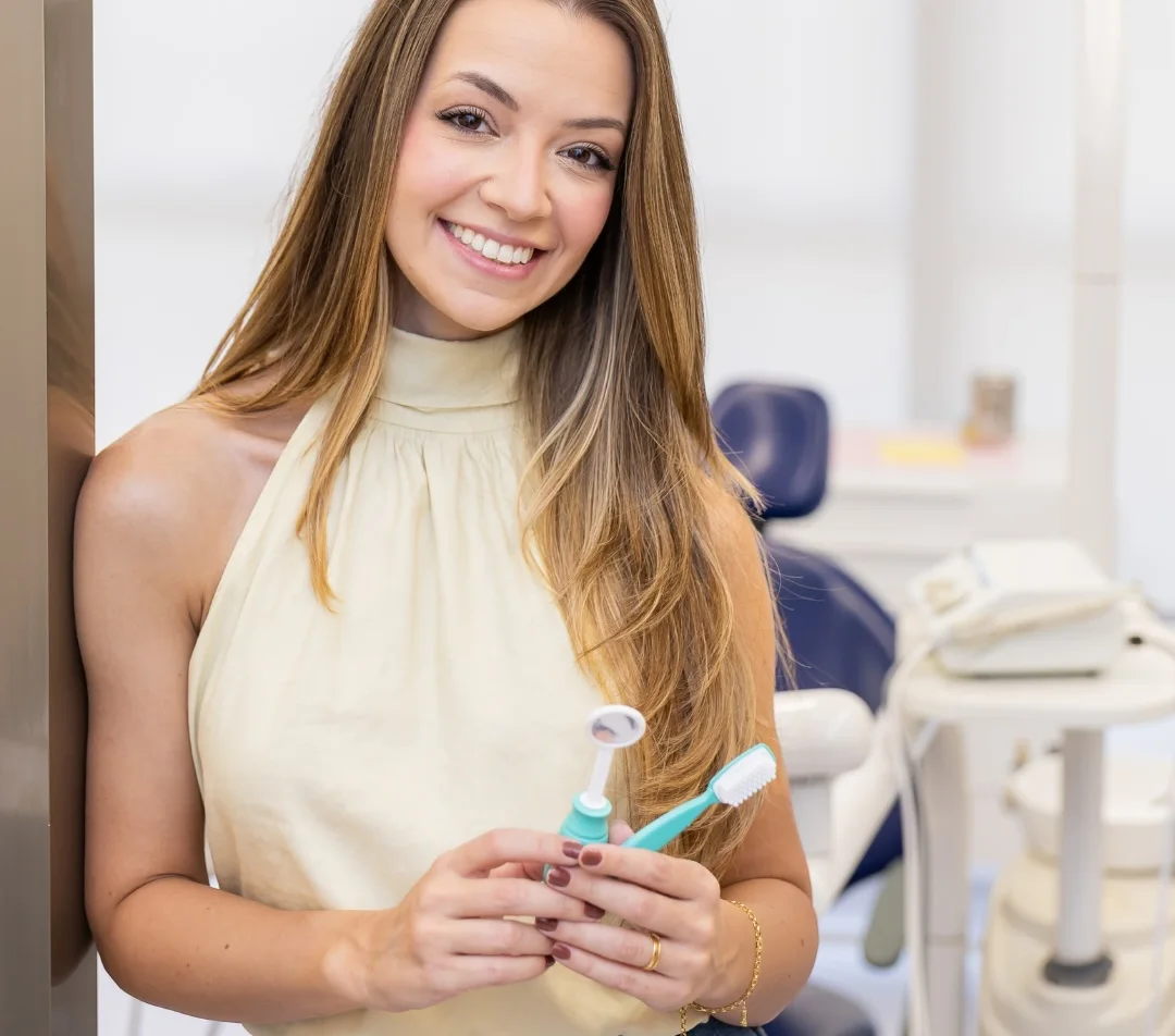Dra. Letícia na sala de atendimento, sorrindo, com uma escova de dentes infantil e um espelho clínico nas mãos