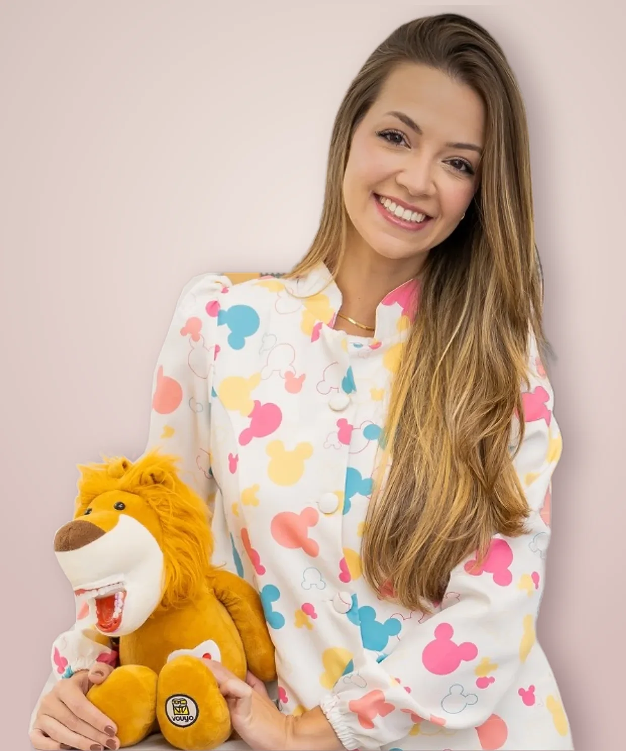Dra. Letícia de jaleco estampado com orelhas do Mickey, abraçada a um leão de pelúcia de boca aberta usado para ensinar escovação