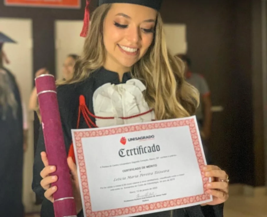 Letícia de beca na formatura, sorrindo, segurando o certificado de mérito do UNISAGRADO