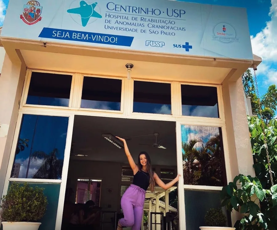 Letícia na porta do Centrinho, o Hospital de Reabilitação de Anomalias Craniofaciais da USP, com o braço erguido sob a placa de boas-vindas