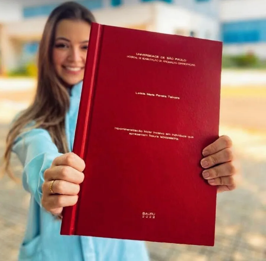Letícia sorrindo e mostrando à câmera o TCC de capa vermelha, de 2022