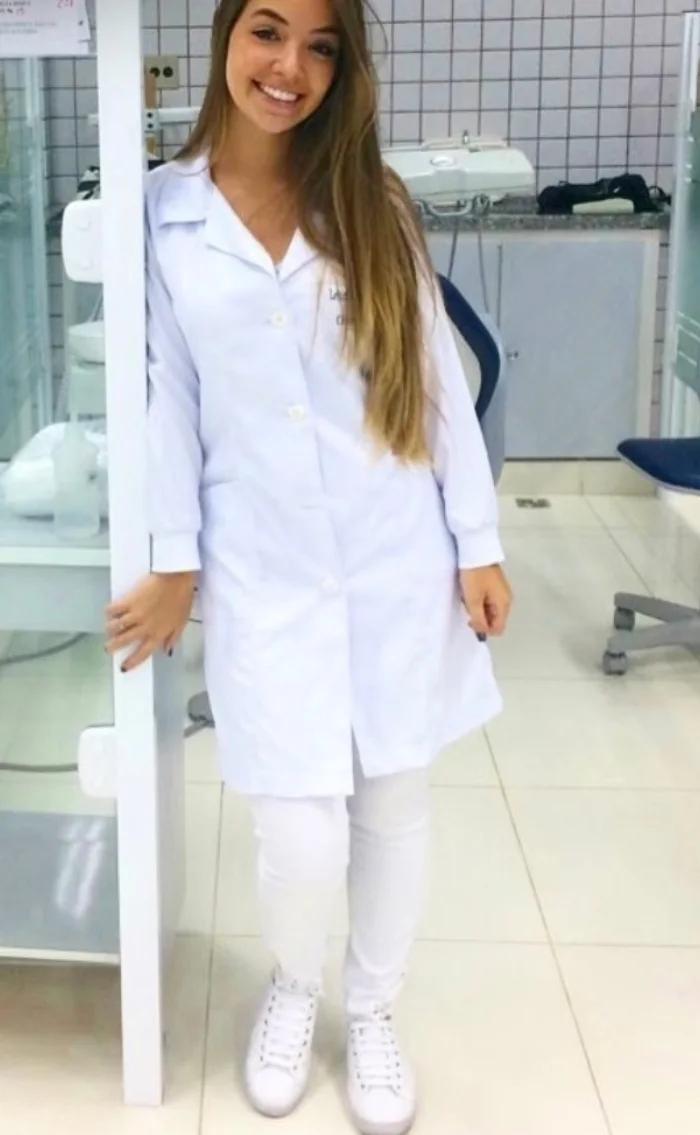 Letícia estudante, de jaleco e calça brancos, de pé na clínica da faculdade
