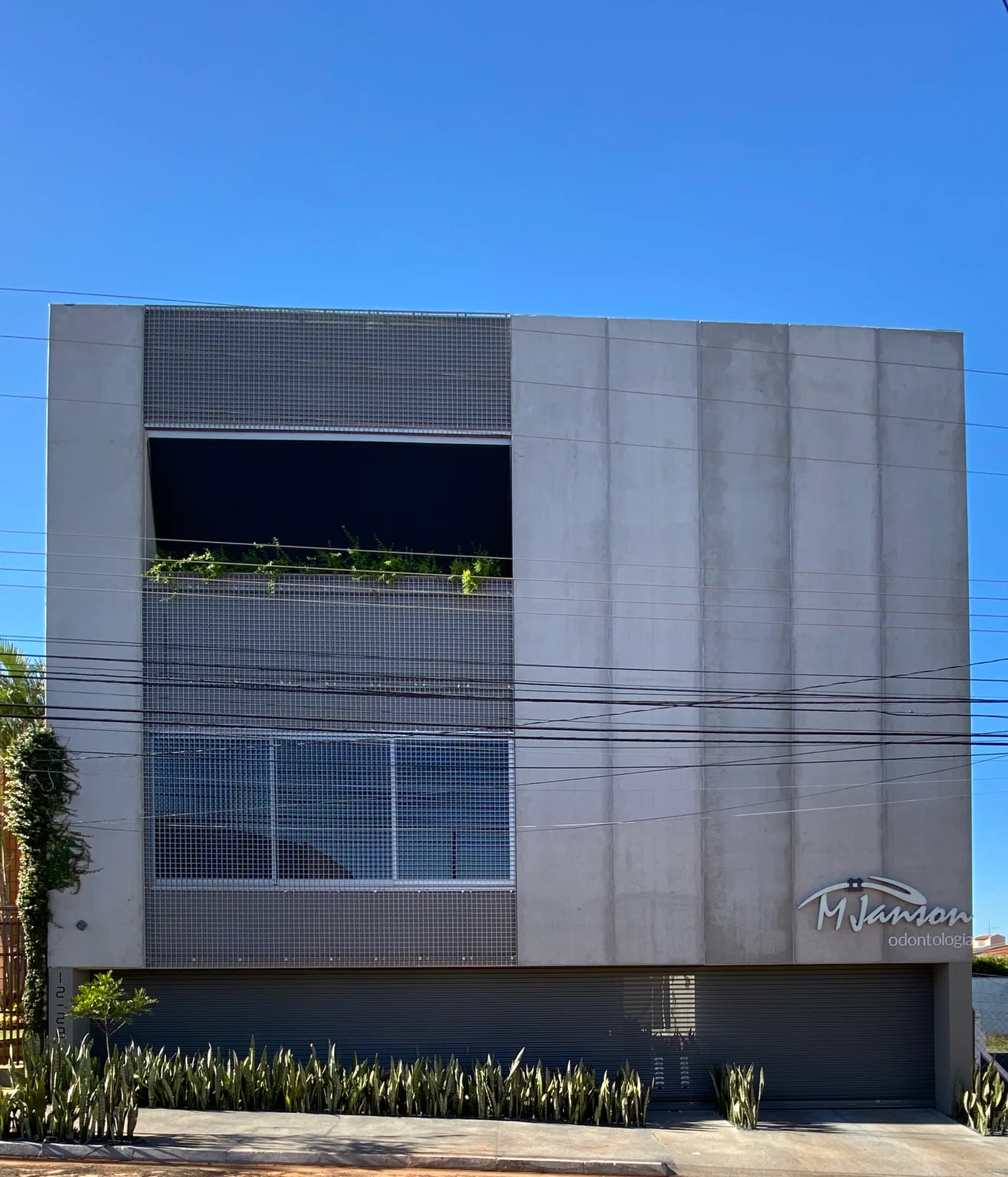Fachada da Clínica MJanson de dia: prédio de concreto aparente, com tela metálica, jardineira e o letreiro MJanson odontologia