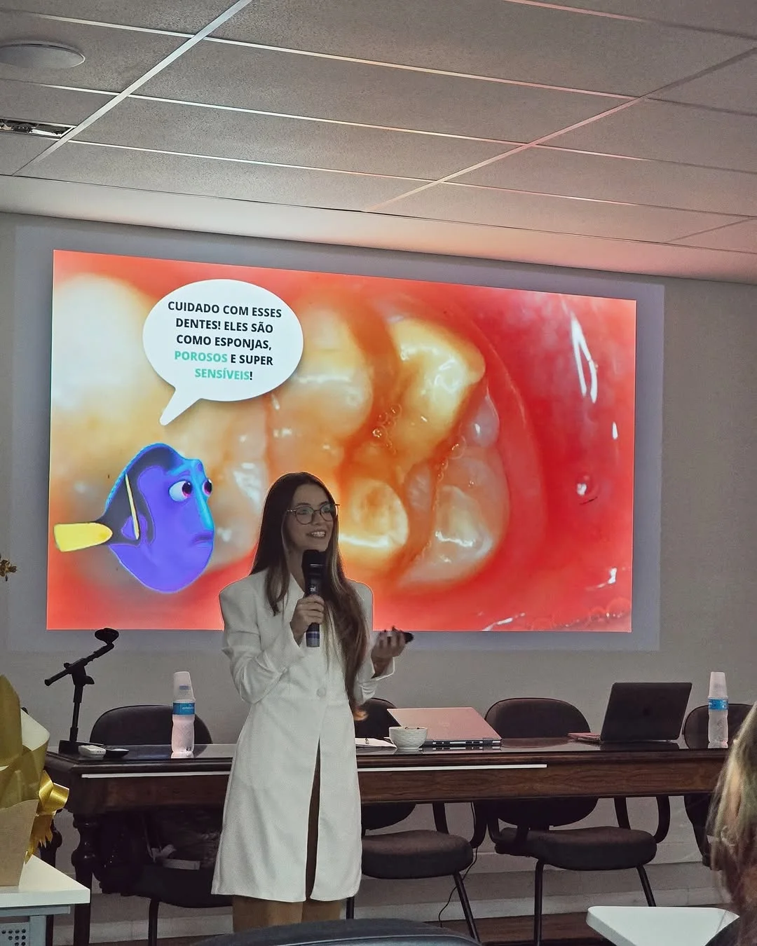 Letícia apresentando com microfone; no telão, foto ampliada de dentes e o balão “Cuidado com esses dentes! Eles são como esponjas, porosos e super sensíveis!”