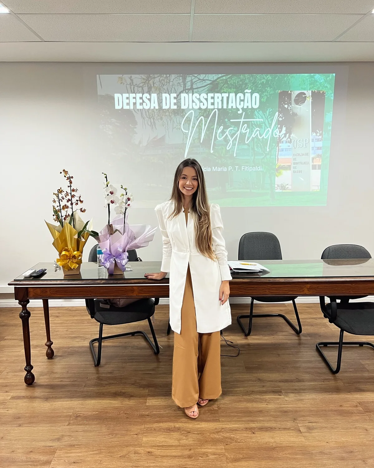 Letícia de blazer branco diante do telão “Defesa de Dissertação · Mestrado”, numa sala da FOB-USP