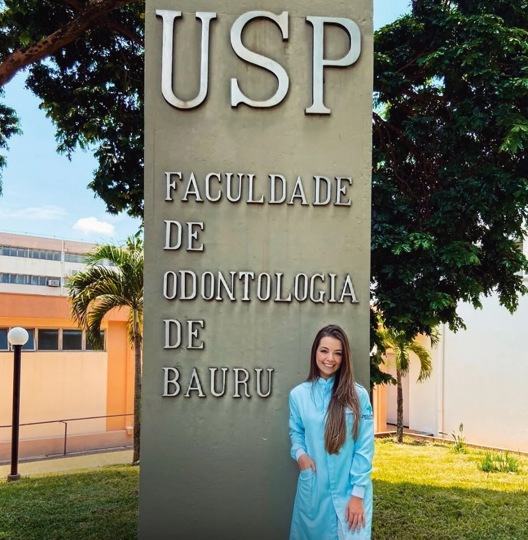 Letícia de jaleco azul ao lado da placa “USP · Faculdade de Odontologia de Bauru”